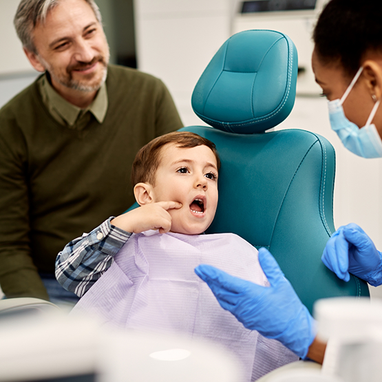 Boy in dental chair pointing to his face while talking to his dentist